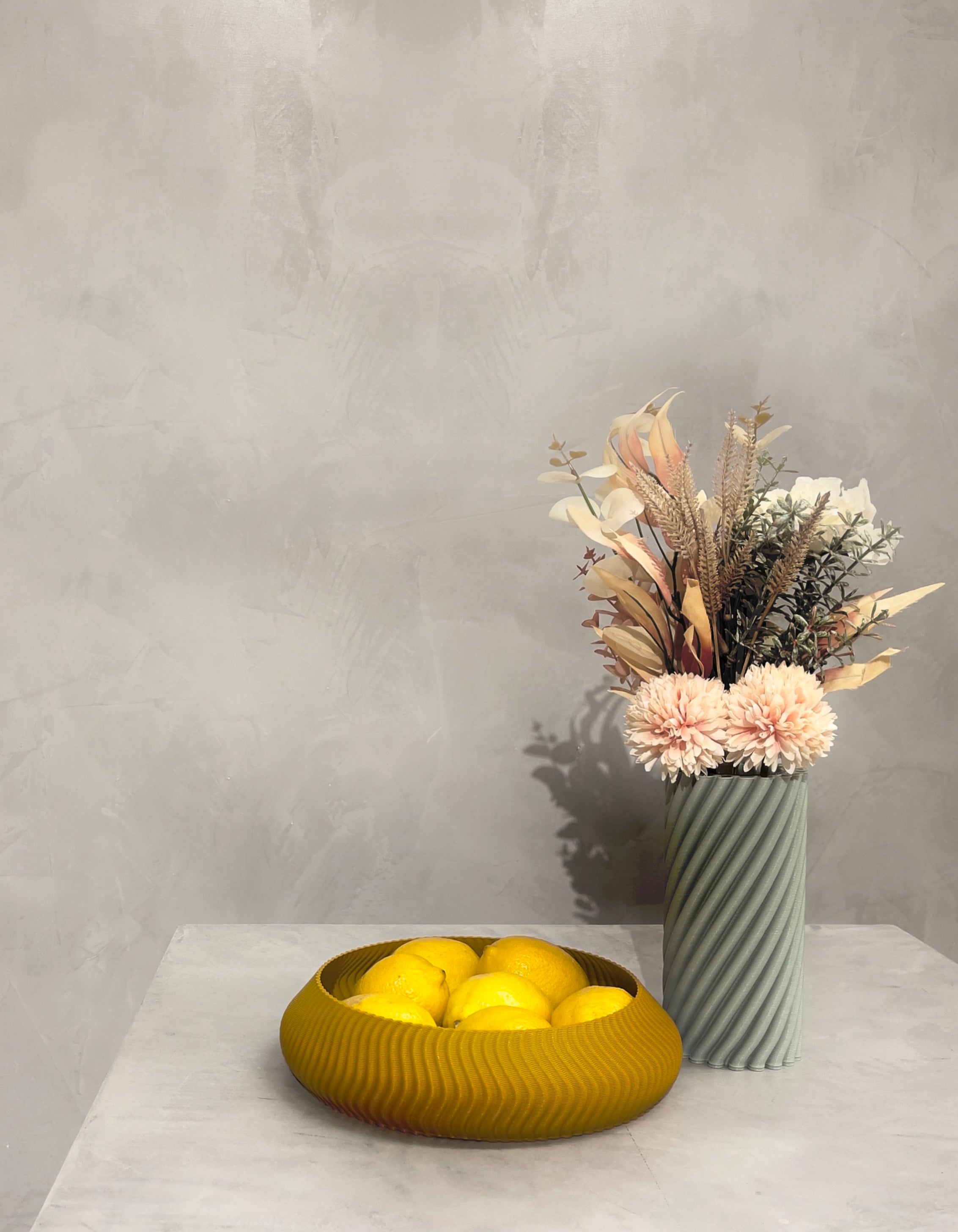 3D printed Kyma bowl with fruit and Strofi vase with flower bouquet made with recycled plastic by Deme Design, pictured on a pedestal